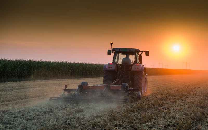 A tractor plowing a field at sunset, with dust rising from the soil, representing modern farming techniques and agricultural machinery.